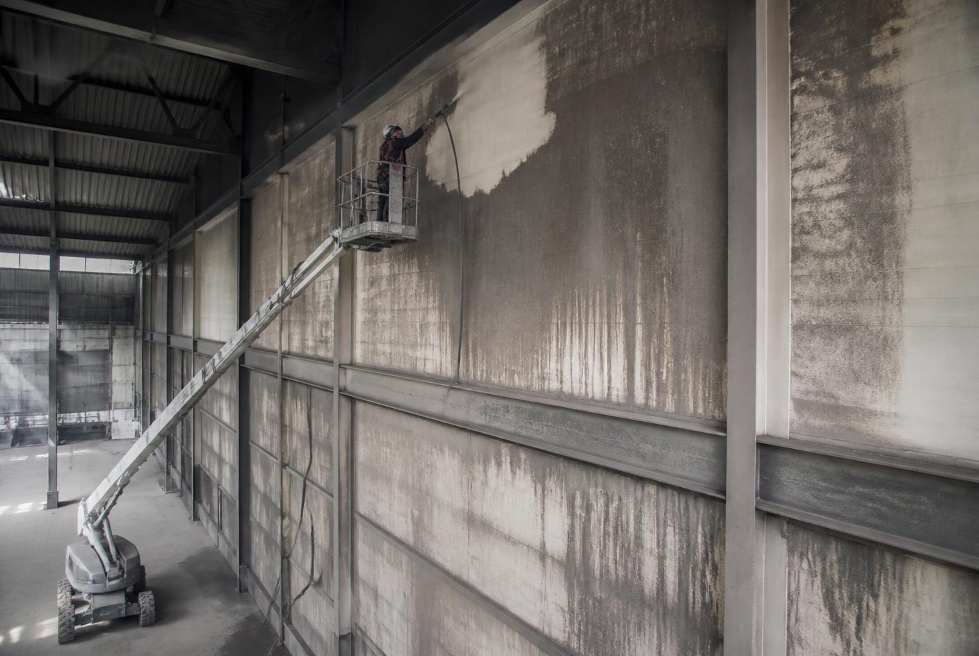Reinigungskraft von Clean Advance in einer Teleskophebebühne, die hohe Hallenwände und Deckenbereiche von Industriestaub mit einem leistungsstarken Industriesauger reinigt